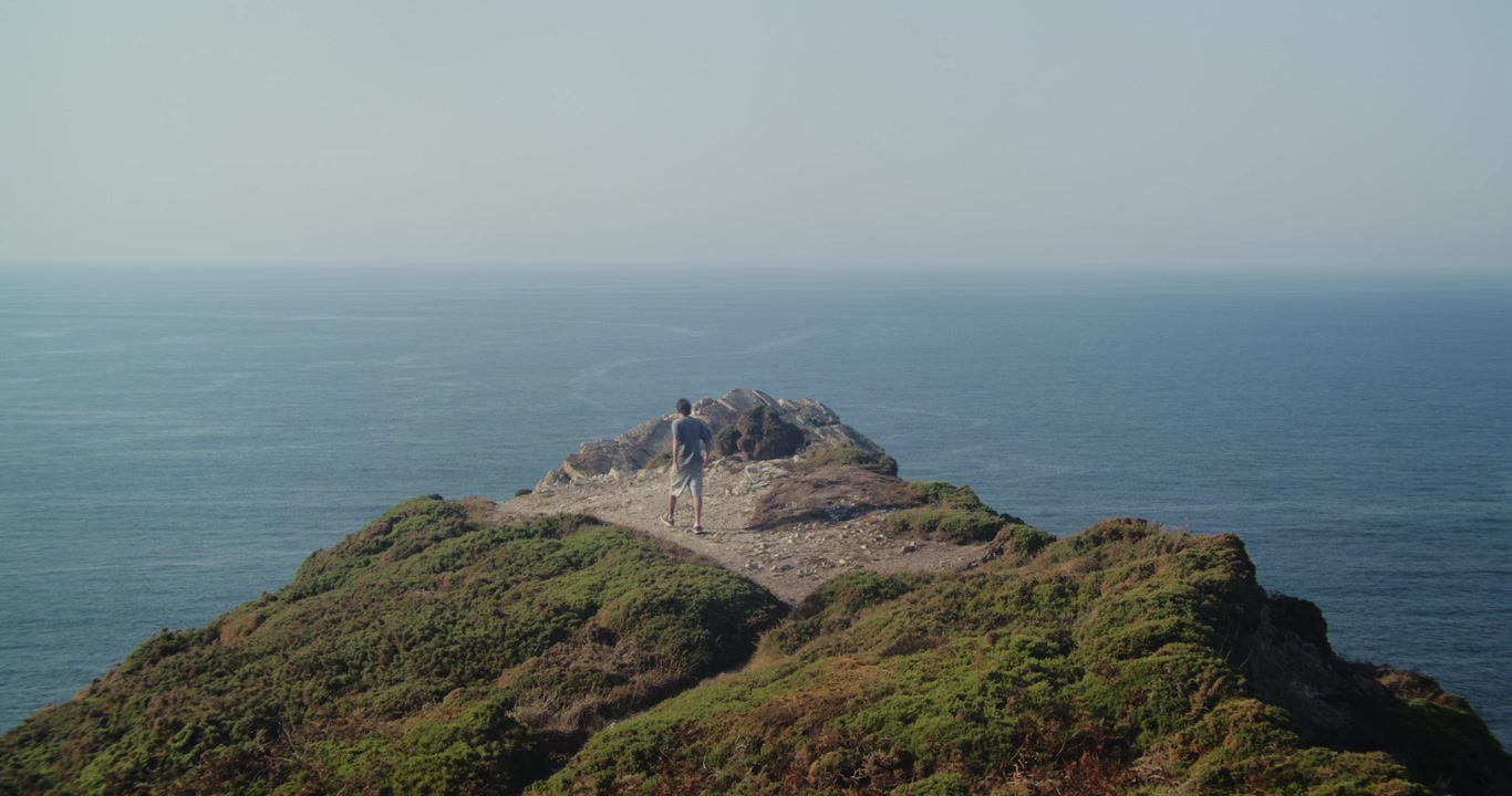 Inquadratura ampia di un uomo che cammina lungo scogliere verdeggianti verso un punto panoramico roccioso sopra l'Atlantico a Cabo Peñas, Asturie. La luce naturale intensa, l'aria costiera e il paesaggio lussureggiante evocano avventura e libertà.