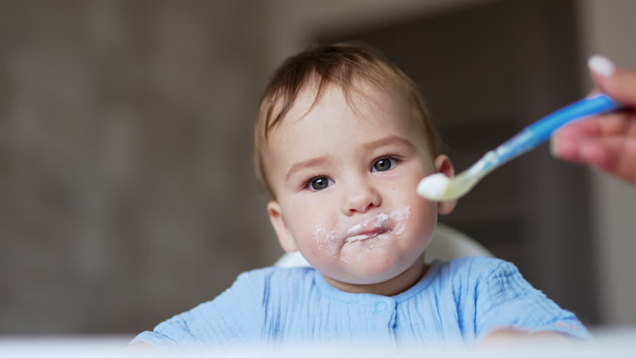 Cute kid eating porridge from spoon. Mother gives porridge to baby and takes the porridge out of kid's face.