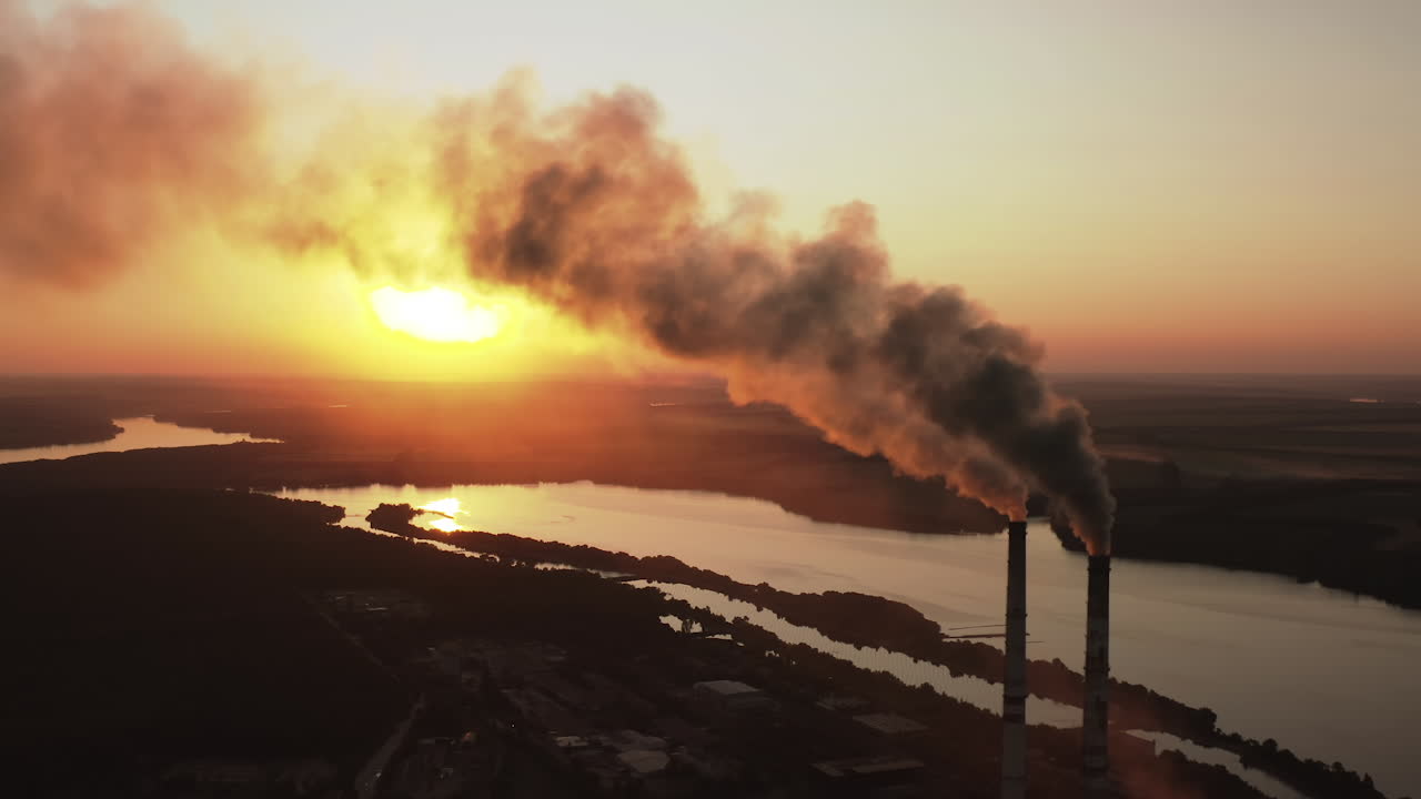 Massive air pollution near the river at darkness. Huge puffs of harmful smoke exhaust into the air against setting sun. Chimneys with dark fumes from manufacture. Aerial view.