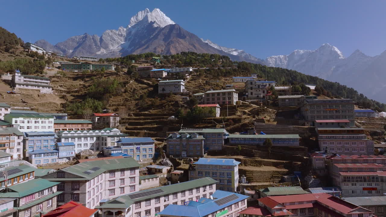 Aerial shot of Namche Bazaar Nepal showing colorful rooftops in an amphitheater-shaped settlement surrounded by green forest, rugged hills and snow-capped Everest Himalayan peaks