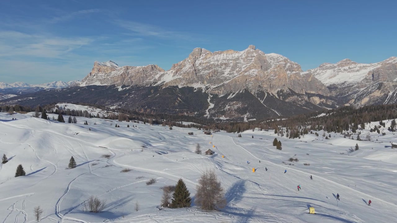 Lyžaři křižující široké zasněžené svahy v Alta Badia pod vrcholy Lavarela a Conturines, se sluncem ozářeným zimním terénem a dramatickými Faneskými Dolomity v pozadí