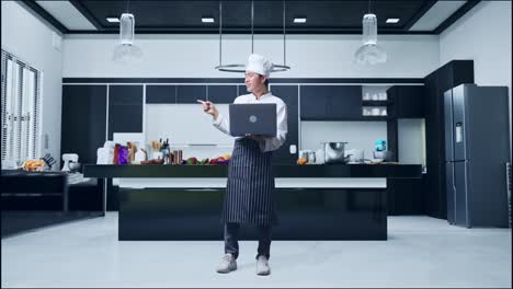 cuerpo lleno de cocinero hombre asiático usando una computadora portátil y apuntando a un lado mientras está de pie en la cocina de casa