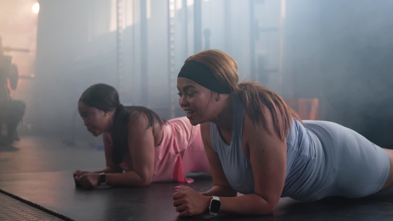 Diverse female friends starting elbow plank on black gym mat, pink, blue tops, watches, motivating