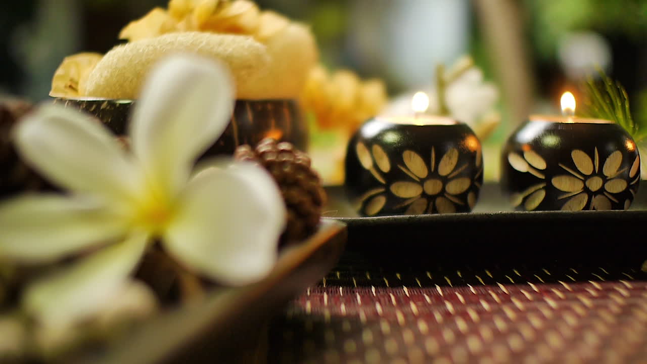 Close‑up of decorative candle holders with floral carvings and lit candles, surrounded by spa elements including plumeria flower, natural textures, and warm relaxing ambiance
