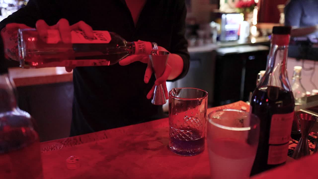 Bartender pours clear liquor into mixing glass under red lighting in a busy bar