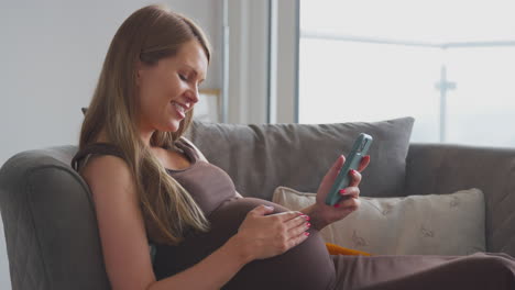 Mujer-Embarazada-Con-Teléfono-Móvil-Relajándose-En-El-Sofá-De-Casa-Sosteniendo-El-Estómago
