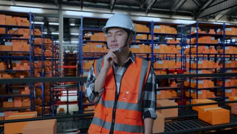 ingeniero masculino asiático con casco de seguridad de pie en el almacén con estantes llenos de bienes de entrega. pensando en algo y mirando a su alrededor en el almacenamiento