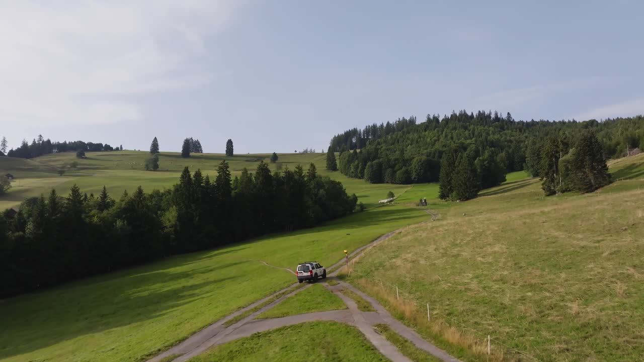 Aerial of a white SUV drives along a winding dirt road through rolling green hills and forested mountains under clear skies, symbolizing freedom, travel, and adventure in nature
