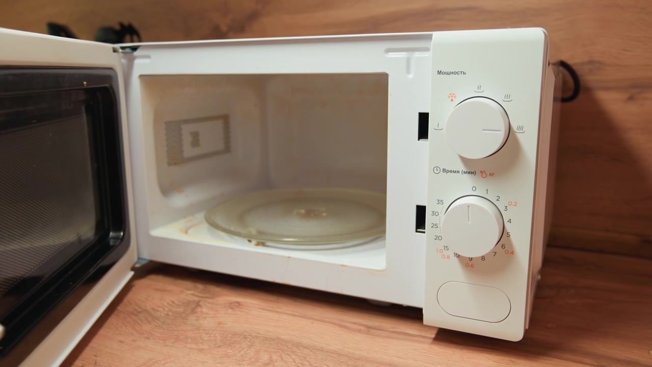Hands in green sleeves open white microwave door and turn lower dial while unit rests on wooden countertop; frontal kitchen view shows empty cavity, glass turntable, soft daylight, domestic task