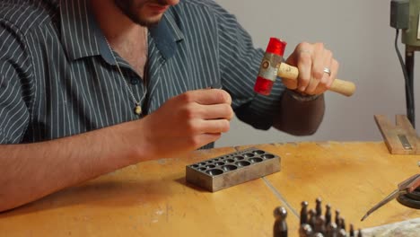 una persona da forma a una pieza de metal usando un martillo y un molde en una mesa de trabajo en un taller bien iluminado, con herramientas en el fondo.