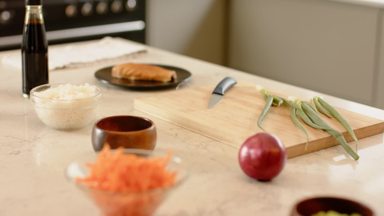 Camera is shifting focus, revealing shredded carrots, rice bowl, soy sauce on marble countertop