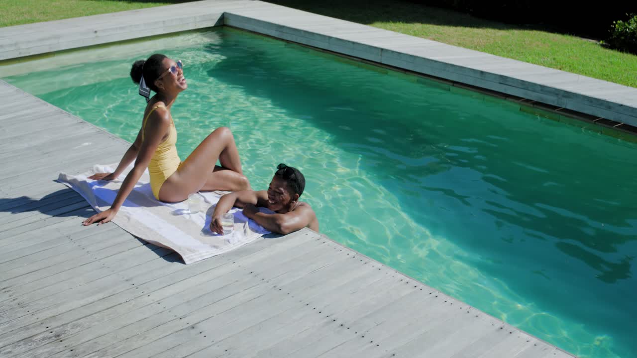 Afroamerikanisches Paar unterhält sich am heimischen Pool im Sonnenschein, während die Frau im gelben Badeanzug sich aus dem Wasser hebt und der Mann lächelt.