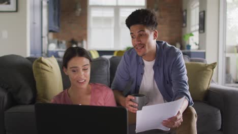 feliz pareja biracial sentada en el sofá de la sala de estar, usando una computadora portátil