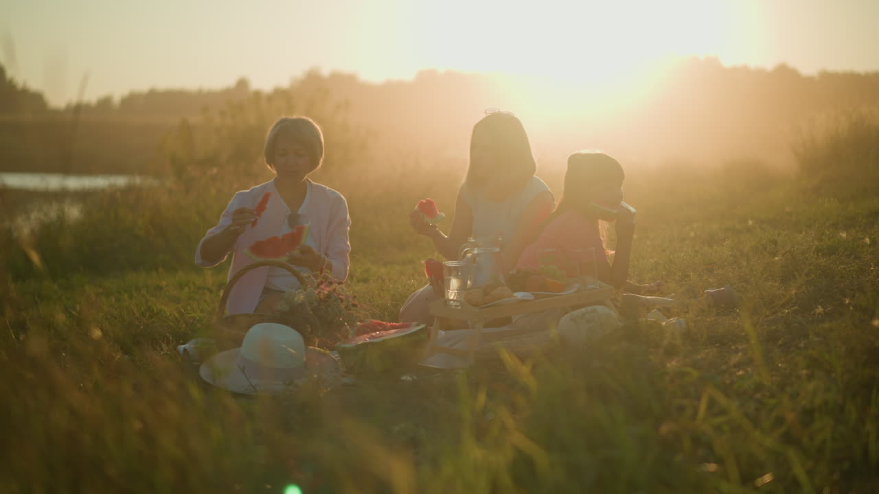 은 오후 피크닉 동안 야외에서 수박을 즐기는 가족, 개방된 잔디에서 휴식을 취하고, 따뜻한 빛은 황금빛을 만들어 평화롭고 즐거운 분위기를 향상시니다.