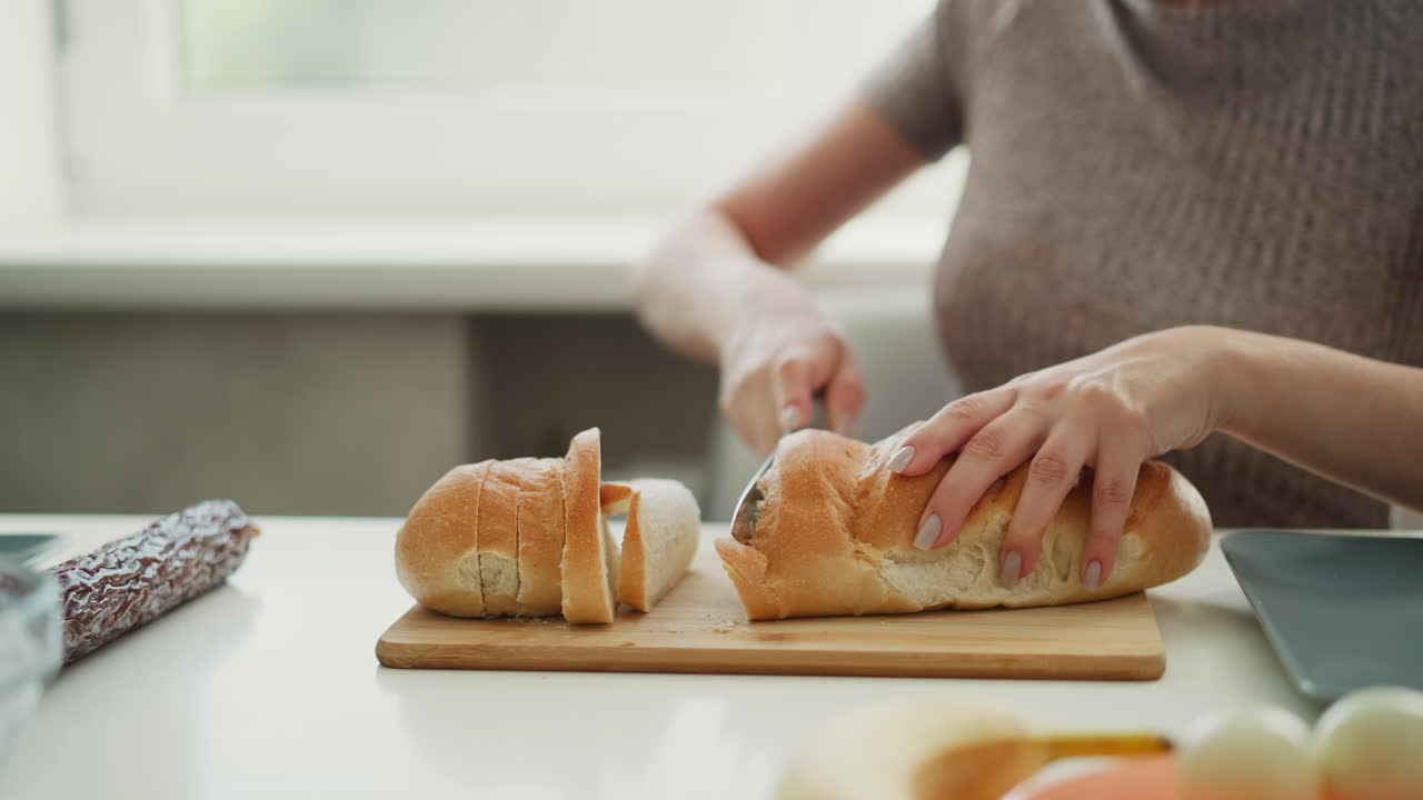 Close up of young woman with manicured nails firmly holding loaf of bread slicing carefully on cutting board next to eggs and plates on kitchen table under natural morning light from window