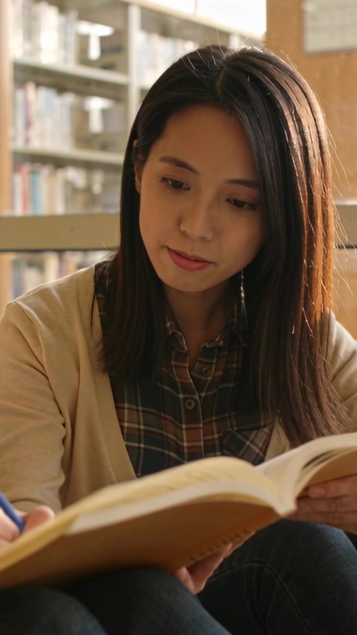 Vertikal video: Asiatisk kvinne ser ned, strekker seg etter en blå penn mens hun leser bok og tar notater.