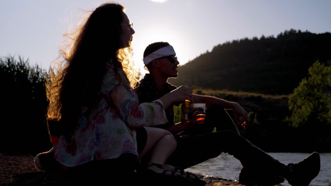 Two friends sitting by a lake, raising drinks in a sunset toast.