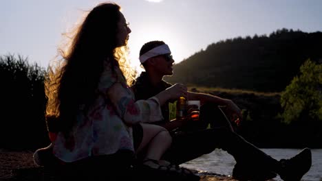 two friends sitting by a lake, raising drinks in a sunset toast.