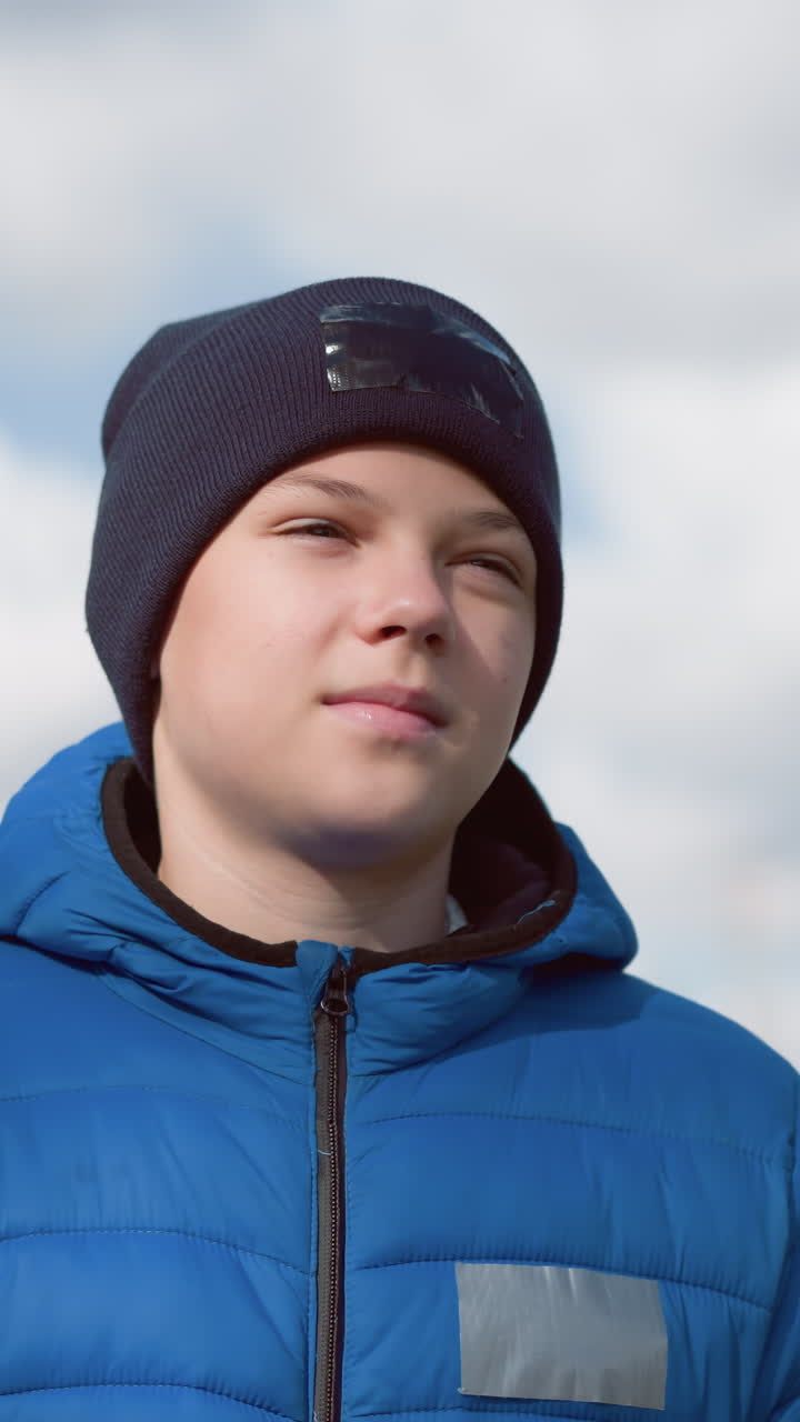 joven con chaqueta azul y gorra negra se pasea por la zona residencial, mirando pensativo con la luz del sol reflejándose en su cara contra los árboles y casas borrosos de otoño