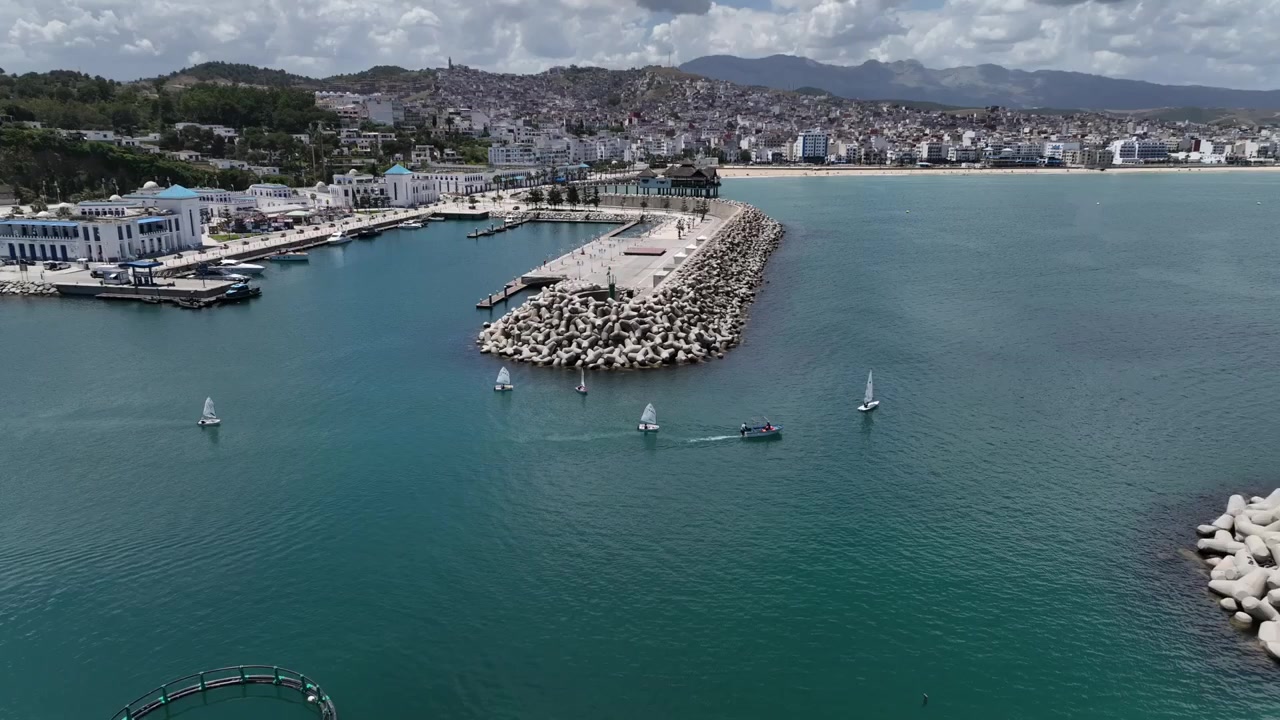 Vista aérea ampla de um porto mediterrâneo protegido no Norte de Marrocos, mostrando múltiplos quebra-mares, água azul calma, pequenos barcos e uma cidade costeira densa subindo em direção às montanhas circundantes