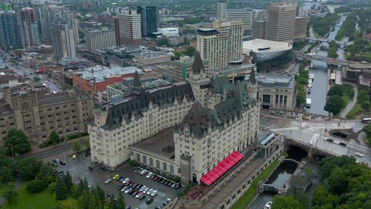 Vista aérea circular suroeste del Fairmont Château Laurier, adyacente a las esclusas del Canal Rideau en un tranquilo día de verano húmedo, con una perspectiva semicenital de dron y otros edificios antiguos en el fondo.