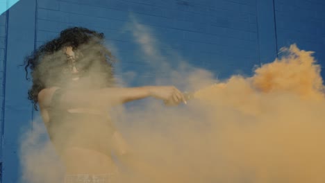 hermosa mujer joven sosteniendo colorida granada de humo bailando afuera contra la pared azul