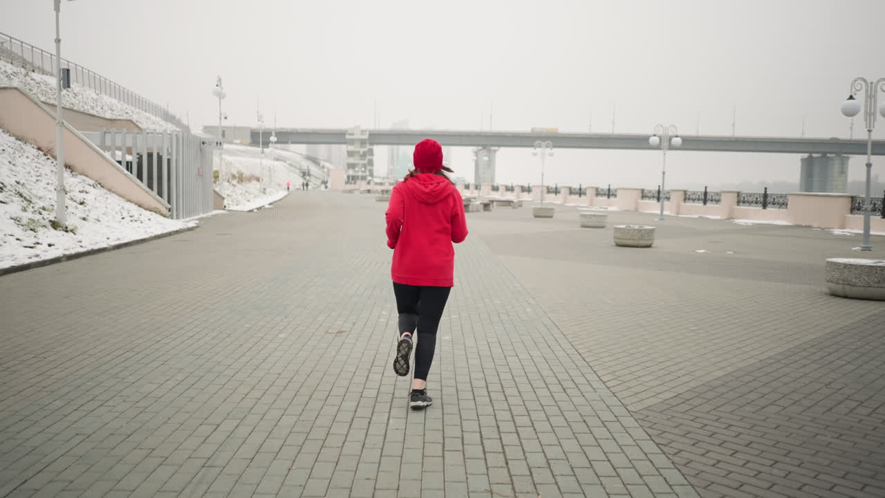 vista trasera de una mujer corriendo al aire libre en el pavimento entrelazado durante el invierno, suelo cubierto de nieve y elementos urbanos modernos, incluidos puentes, postes de luz y bancos