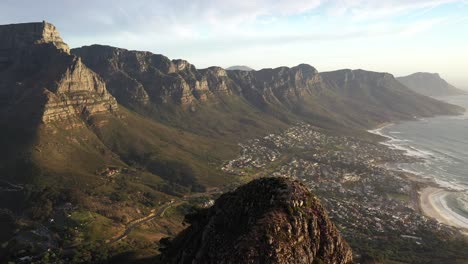 dramatic pull away cinematic aerial shot revealing cape town's lion's head peak with table mountain, camps bay, and clifton during golden hour sunset