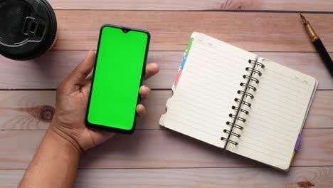 mano sosteniendo un teléfono inteligente con pantalla verde y cuaderno en una mesa de madera