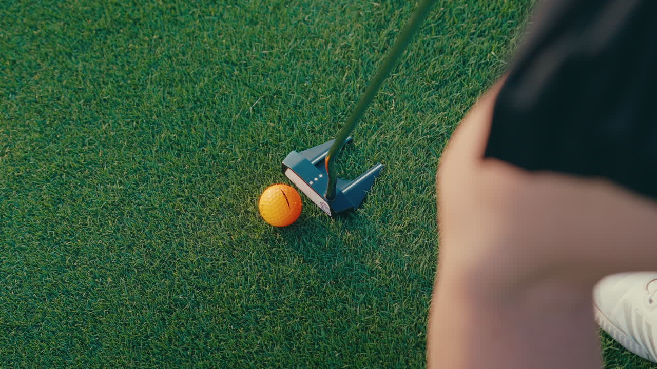 Top-down shot of female golfer putting an orange ball on the green. Her leg, putter, and the ball are visible, capturing a clean overhead view of a focused putting moment in natural light.