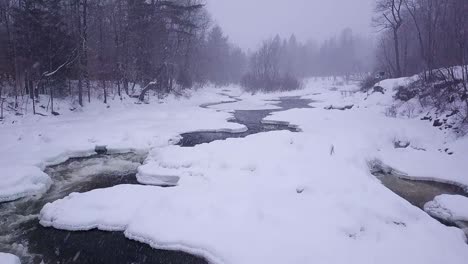 SLOW-MOTION-aerial-shot-flying-further-upstream-along-an-icy-river-during-a-winter-snow-storm