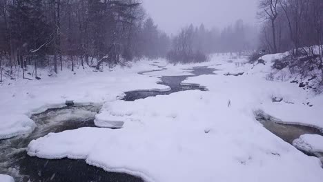 slow motion aerial shot flying further upstream along an icy river during a winter snow storm