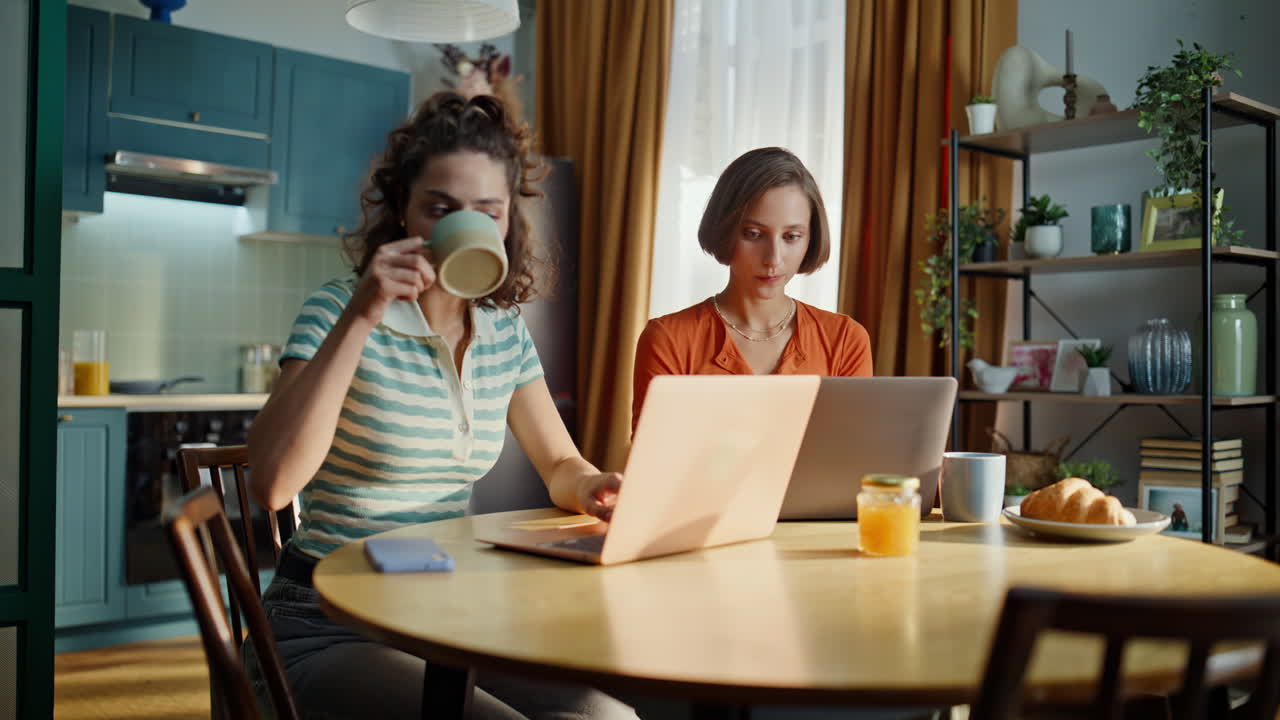 Deux femmes regardant des ordinateurs et prenant leur petit-déjeuner ensemble dans une cuisine moderne. Deux femmes