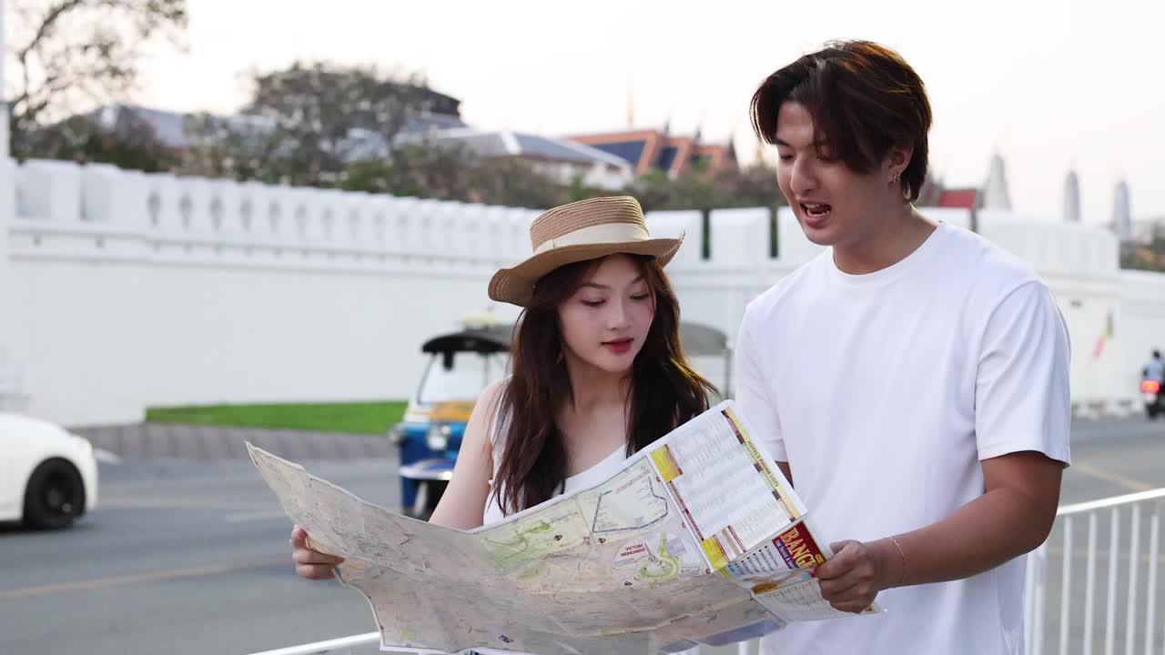 Coppia asiatica consulta una mappa cartacea all'aperto, discutendo indicazioni vicino a un muro bianco storico al tramonto