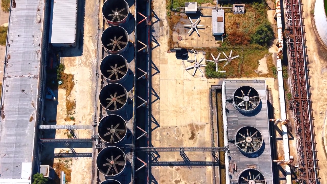 Top down aerial view of industrial cooling fans and vents. Large circular cooling units sit on concrete structures beside metal framing during bright daytime.