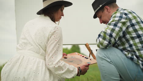 a man and woman are bent over in a grassy field, preparing an outdoor painting session. the woman in a white dress holds a wooden palette, while the man in a checked shirt carefully squeezes out paint