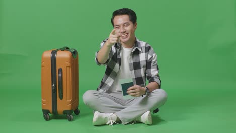 full body of asian male traveler with luggage and passport smiling and touching his chest then pointing at you while sitting in the green screen background studio