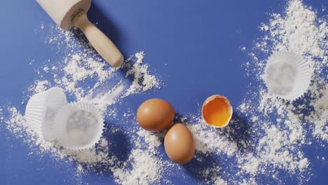 video of baking ingredients, muffin papers, eggs and tools lying on blue surface with flour