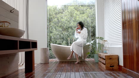 biracial woman wearing bathrobe sitting on bathtub drinking tea in bathroom at home, slow motion