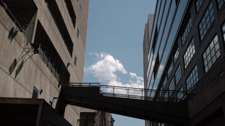 Vista de baixo para cima de um moderno trem de metrô branco atravessando uma passarela elevada que conecta um edifício de concreto a um com fachada de vidro, tudo sob um belo céu azul claro com nuvens fofas.