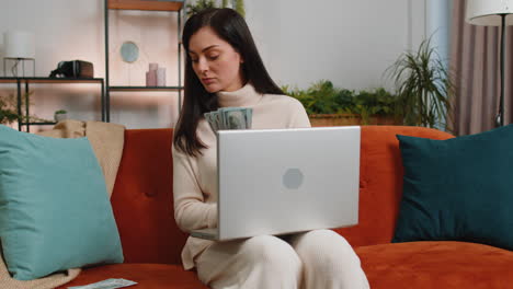 Una-Mujer-Feliz-Y-Sonriente-Contando-Dinero-En-Efectivo-Y-Usando-Una-Computadora-Portátil-Para-Calcular-Los-Ingresos-Domésticos-En-Casa