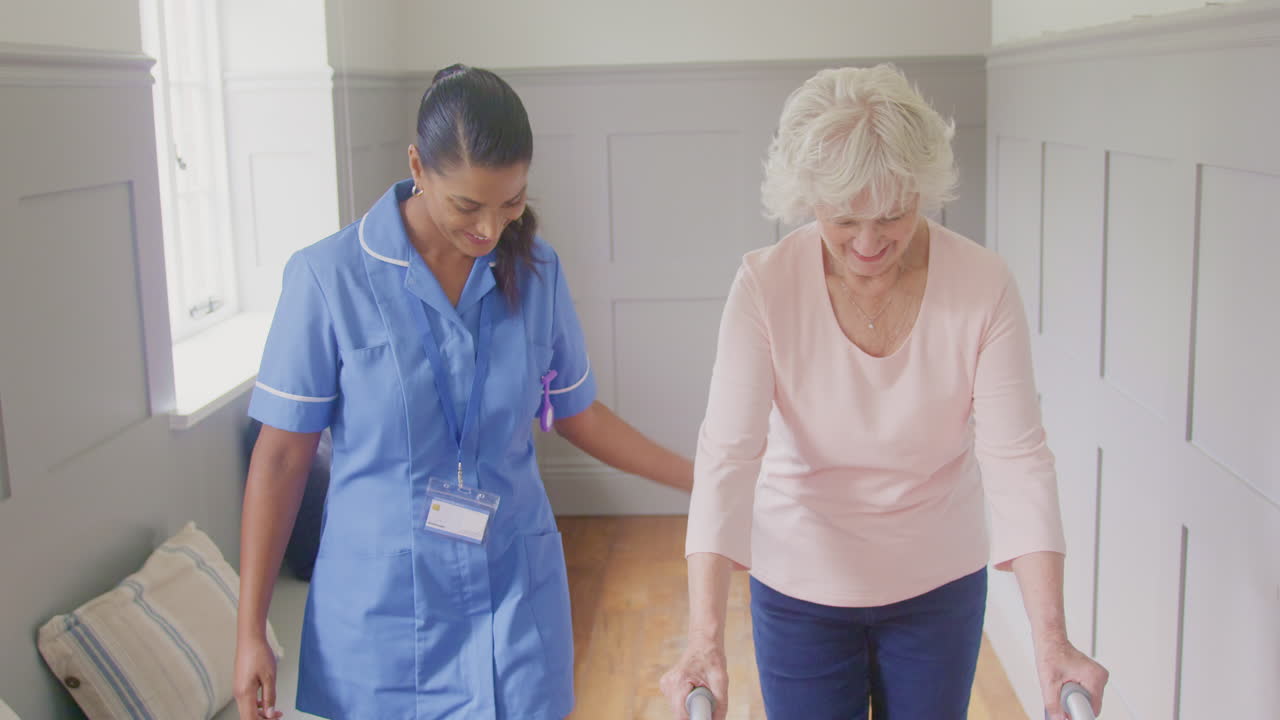 Senior Woman At Home Using Walking Frame Being Helped By Female Care Worker In Uniform