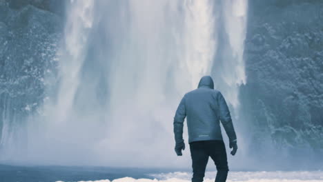 young traveler in blue jacket with huge frozen waterfall in the background in slow motion