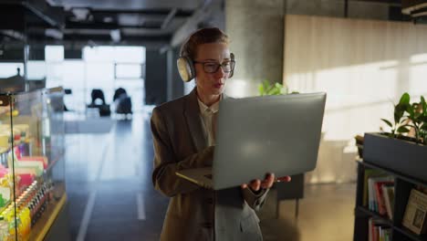 A-confident-middle-aged-blonde-girl-with-glasses-in-a-gray-business-suit-walks-through-a-modern-office-and-types-on-her-laptop-while-working-in-a-sunny-office