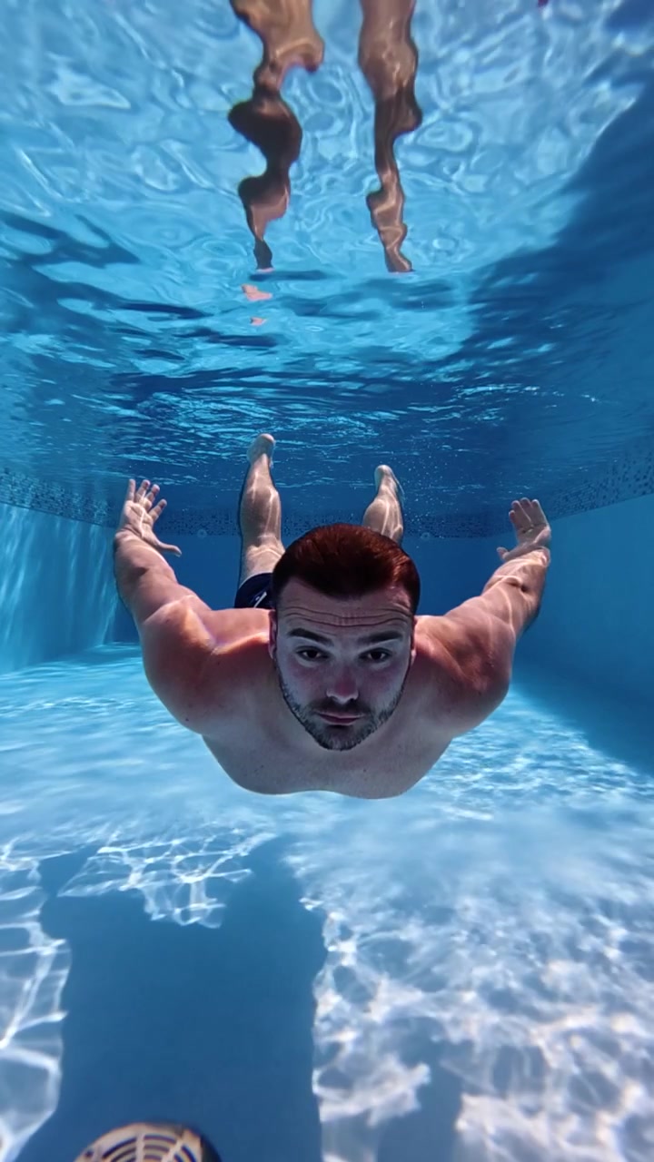 Vista subacquea di un uomo che si tuffa e nuota in una piscina blu limpida, ripreso in super slow motion a 240 fps con bellissimi riflessi di luce
