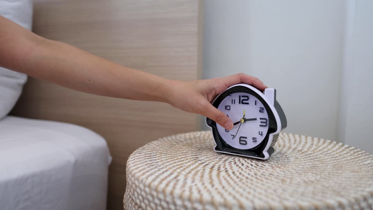 Una mano si allunga dal letto per afferrare una sveglia nera su un comodino.