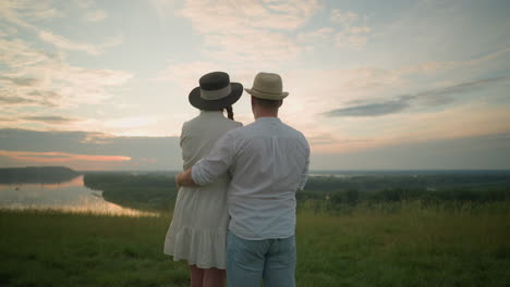 a close-up of a couple standing on a grassy hill at sunset, gazing at the sky. the man, in a white shirt, hat, and jeans, gently holds the woman by the waist. she wears a black hat and a white dress