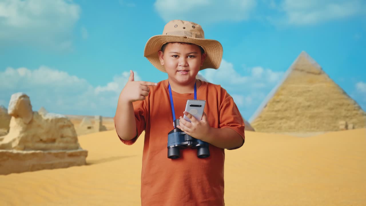Asian Boy With A Hat And Binoculars Smiling And Pointing To Smartphone While Traveling In Giza Pyramid. Boy Researcher Examines Something, Travel Tourism Adventure Concept