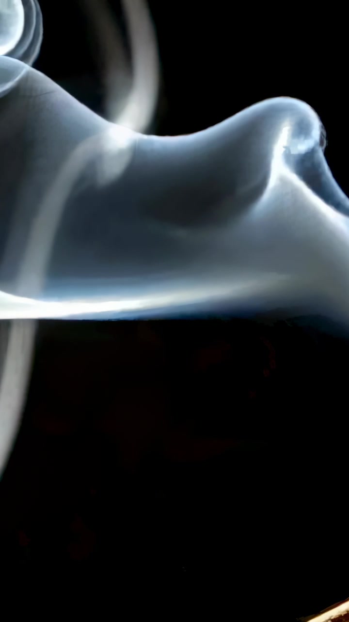 Vertical video: Rising bluish smoke ribbons curling into spirals on black backdrop, with ember glow