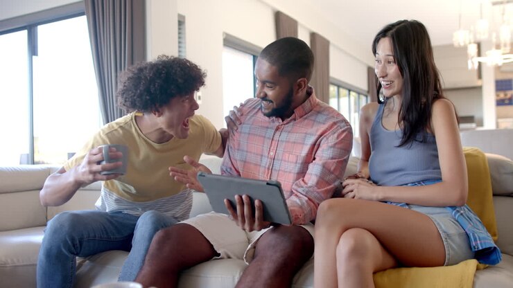 At home, diverse friends hanging out, using tablet and celebrating while sitting on couch